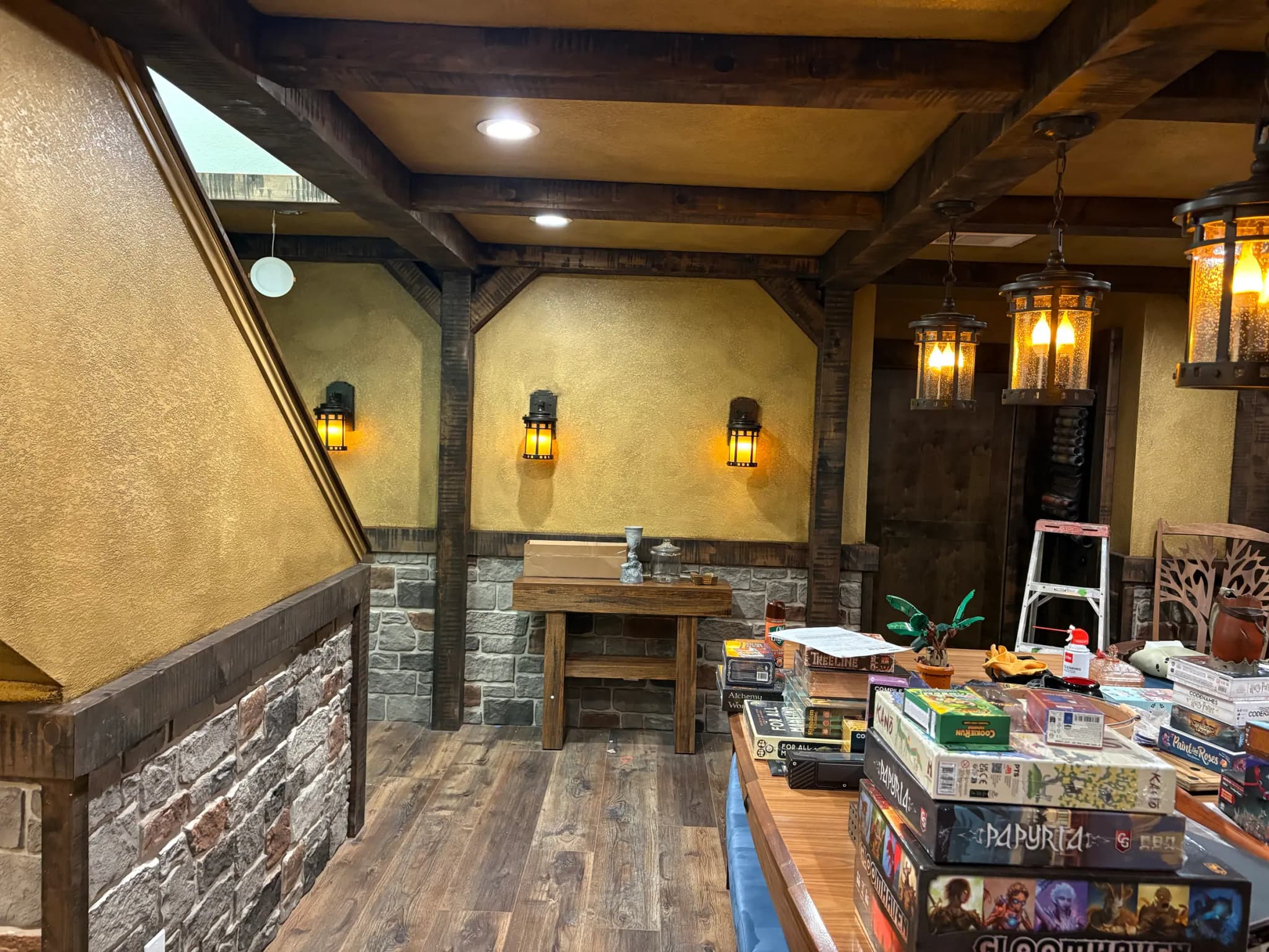 A game room finished like a tavern: heavy timber beams and posts, textured ochre walls over a stone wainscot, lantern pendants and a plank floor.
