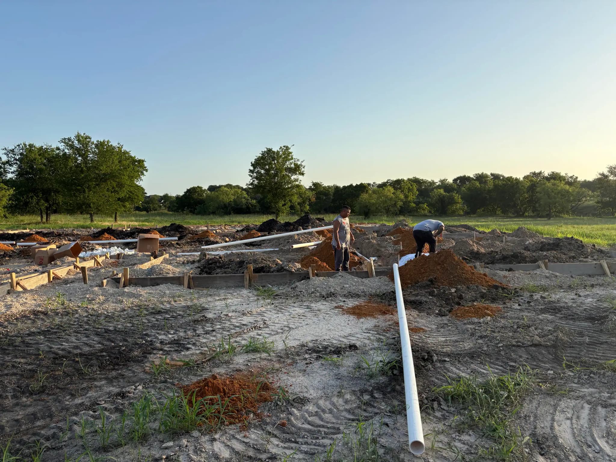 Footing forms and grade-beam trenches across a cleared pad, spoil piles either side, a length of white drain pipe laid across the foreground and two people working at the far trench.