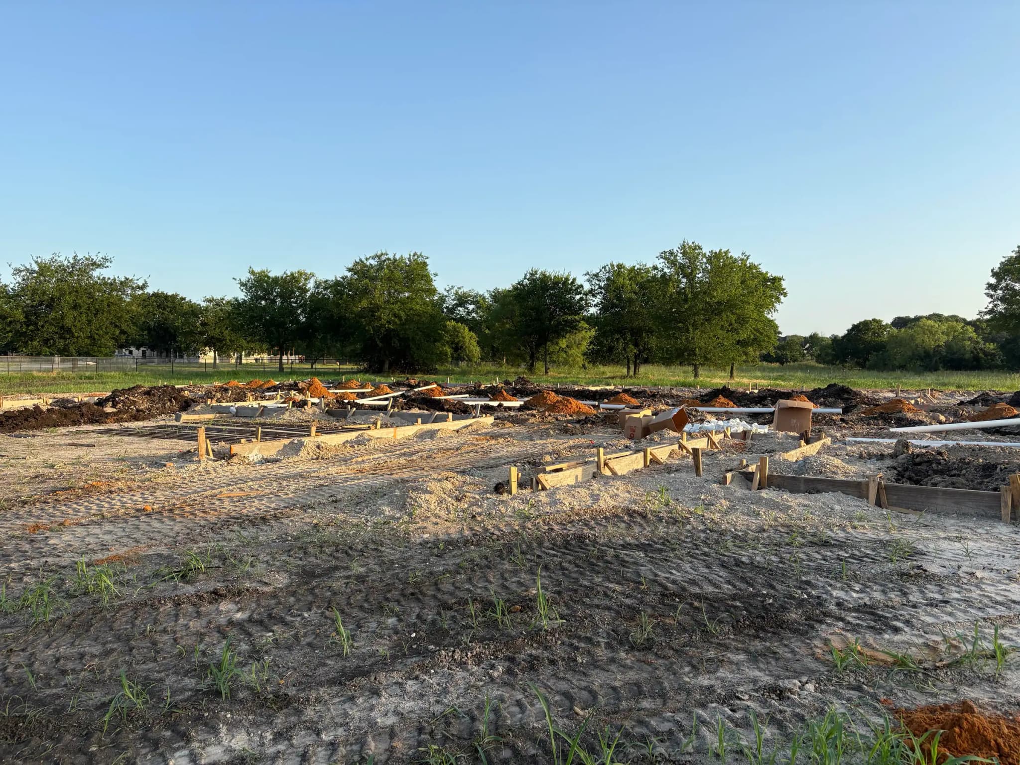 A cleared building pad with footing forms and reinforcing set out in a grid, trenches cut for grade beams and tracked-machine marks across the sand.