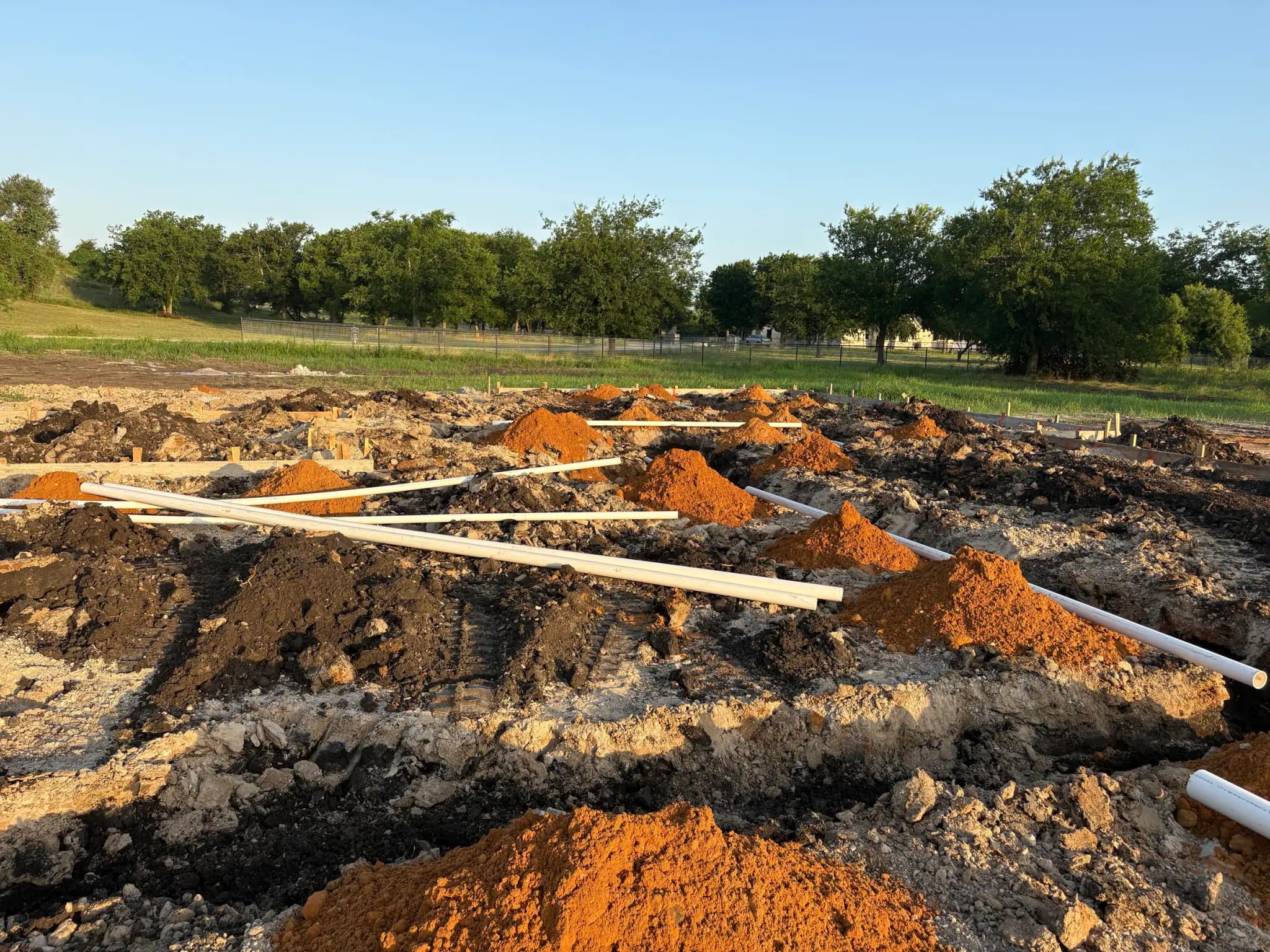 Lengths of white drain pipe stacked across mounded orange and gray spoil on an excavated pad.