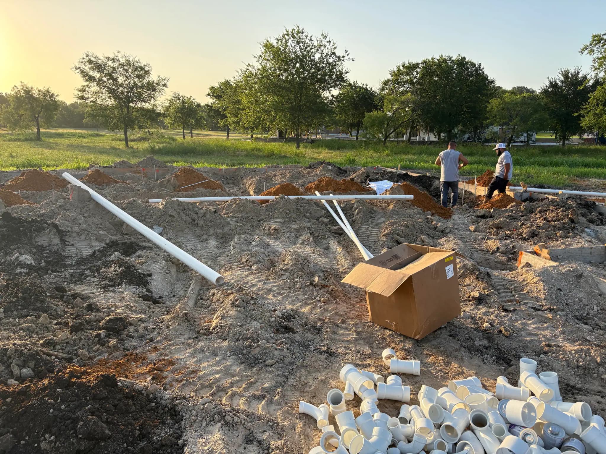 Drain runs laid over an excavated pad in low light, a carton and a pile of white pipe fittings in the foreground and two people at the trench beyond.