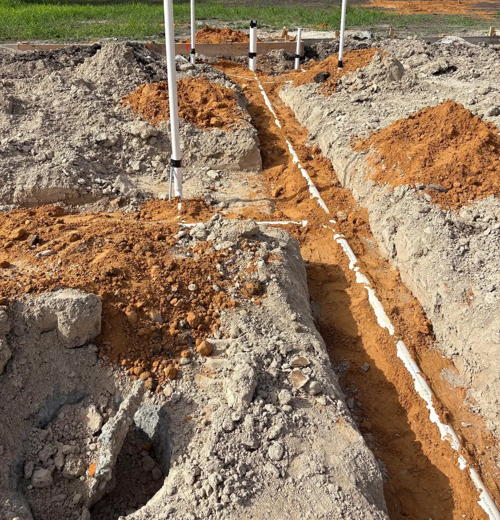 A narrow trench with white drain pipe bedded along its floor, a riser standing at a junction and formwork behind.