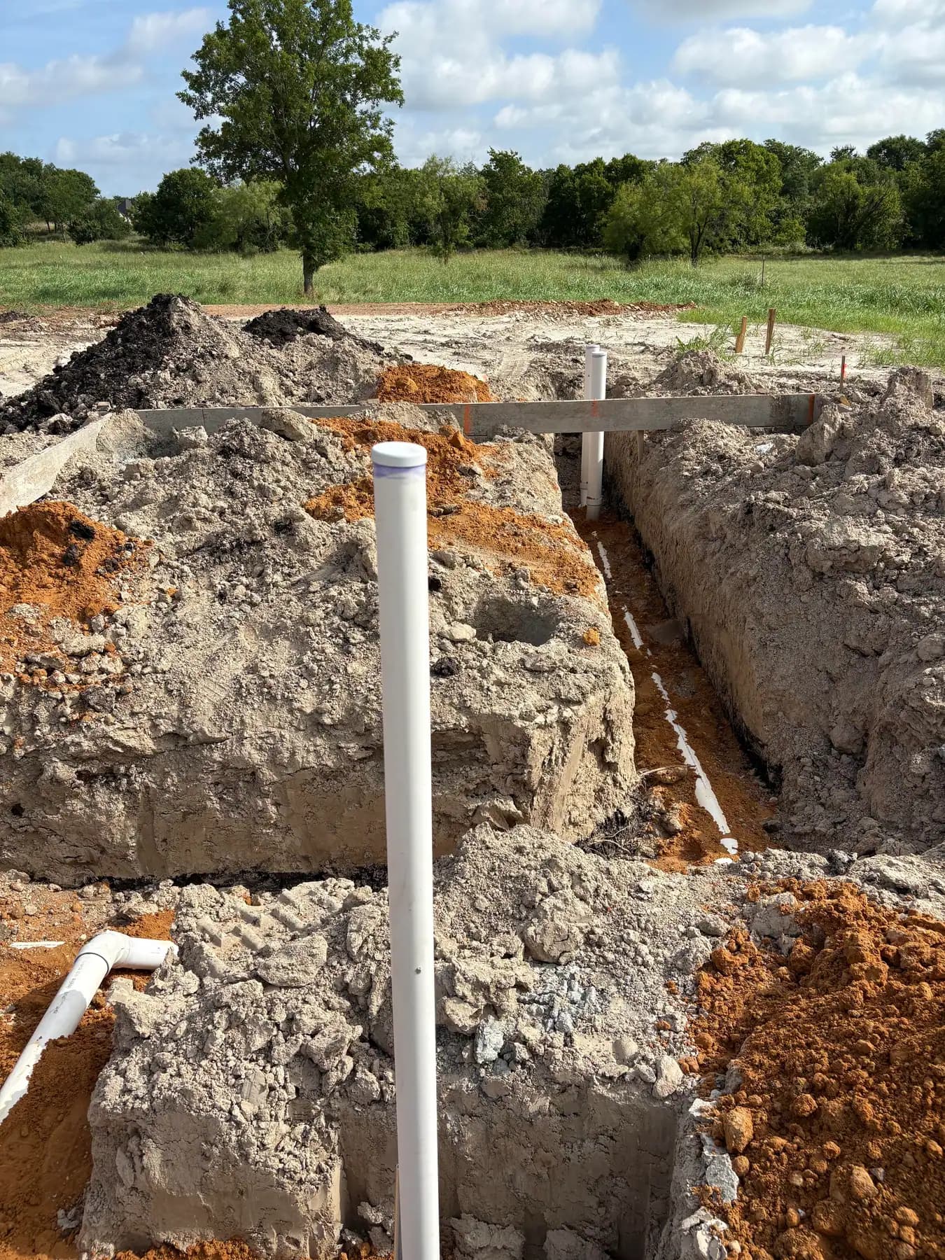 A white pipe riser standing in an open trench between banks of excavated pale rock and orange clay.