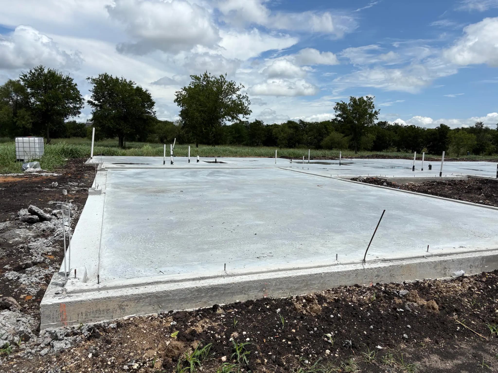 A finished concrete slab poured across the whole footprint, plumbing stub-ups standing through it, seen from one corner under a cloudy sky.