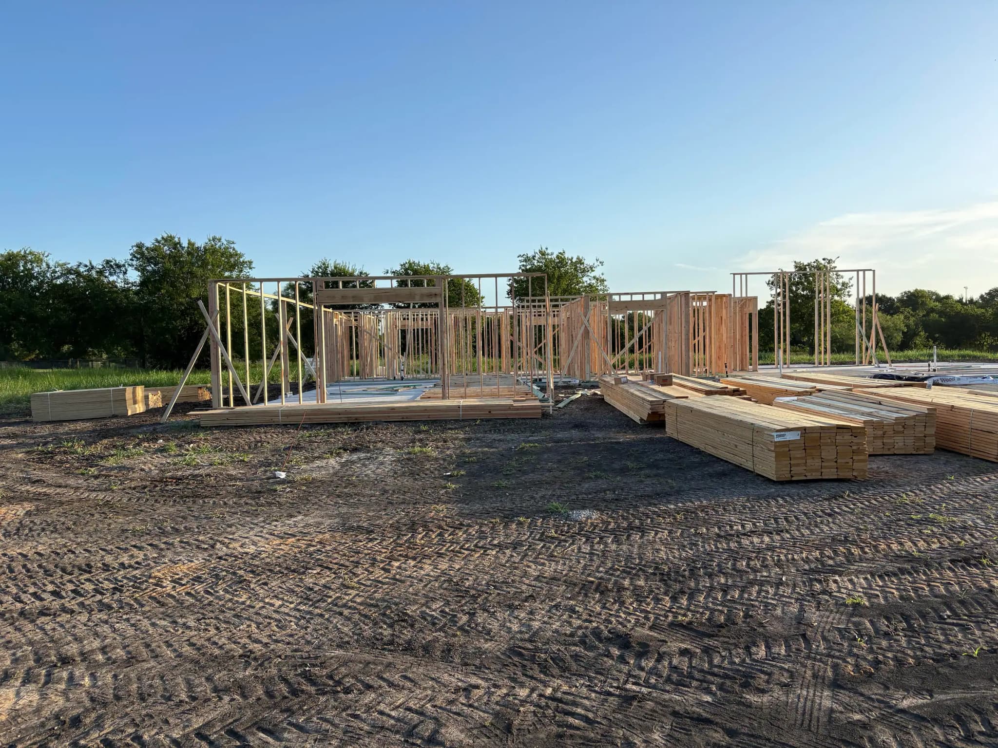 Exterior stud walls standing on the slab with temporary bracing, and banded packs of framing lumber stacked on the ground in front.