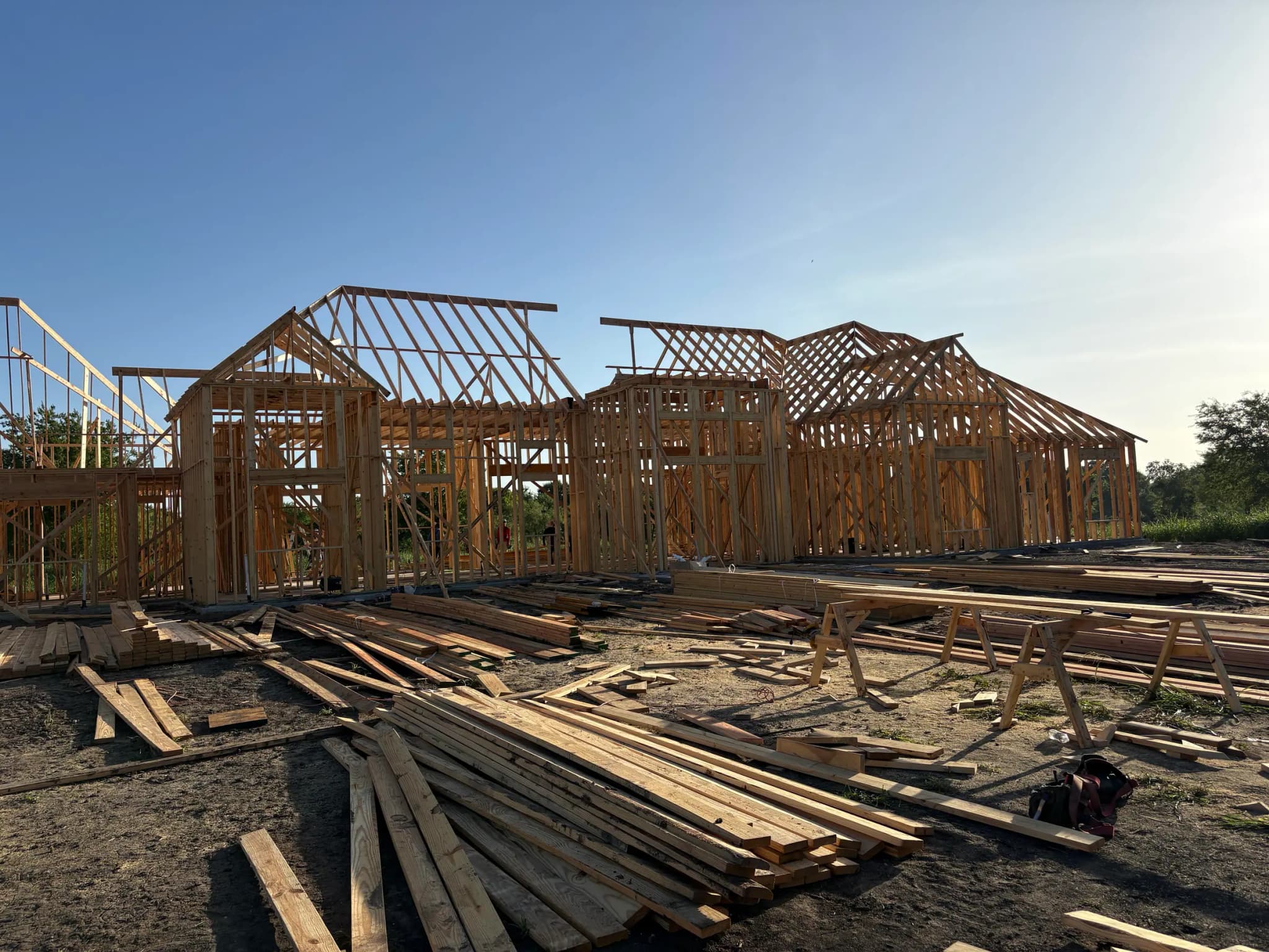 Roof trusses set over two gables of the framed house, with cut offcuts and sawhorses on the ground below.