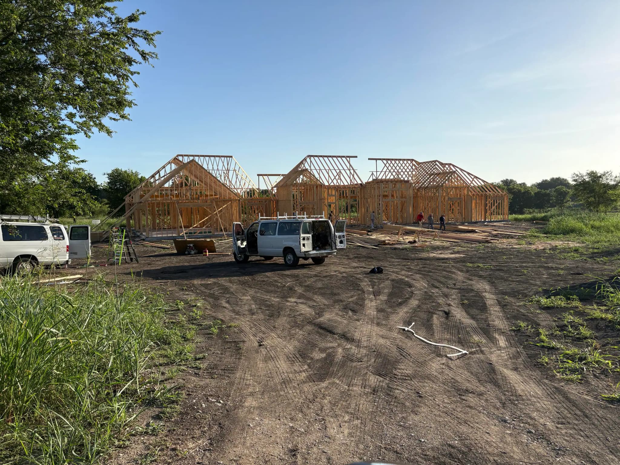 The framed house with roof trusses across three gables, work vans parked on the graded drive and several people working under the frame.