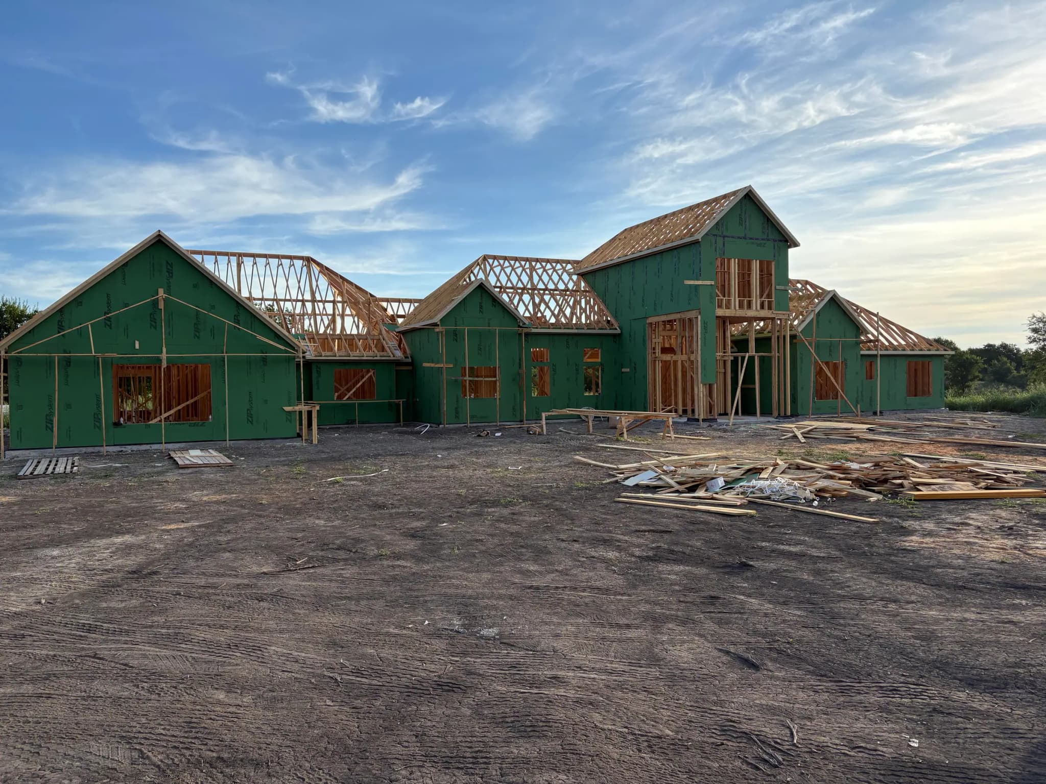 The whole elevation sheathed in green wall panels with roof trusses set across four gables, offcuts stacked on the graded ground in front.