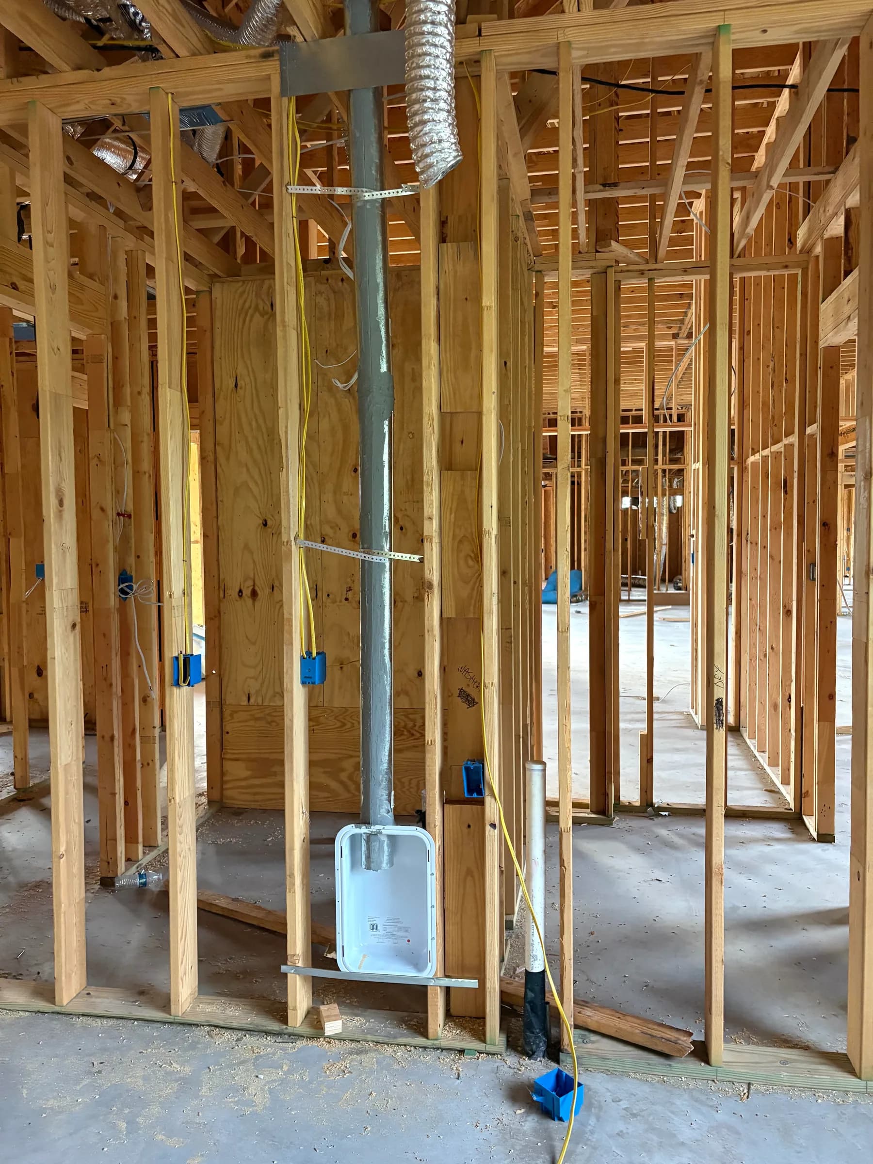 An interior stud wall with a metal flue rising through it, yellow electrical cable stapled to the studs and rough-in boxes set at switch height.