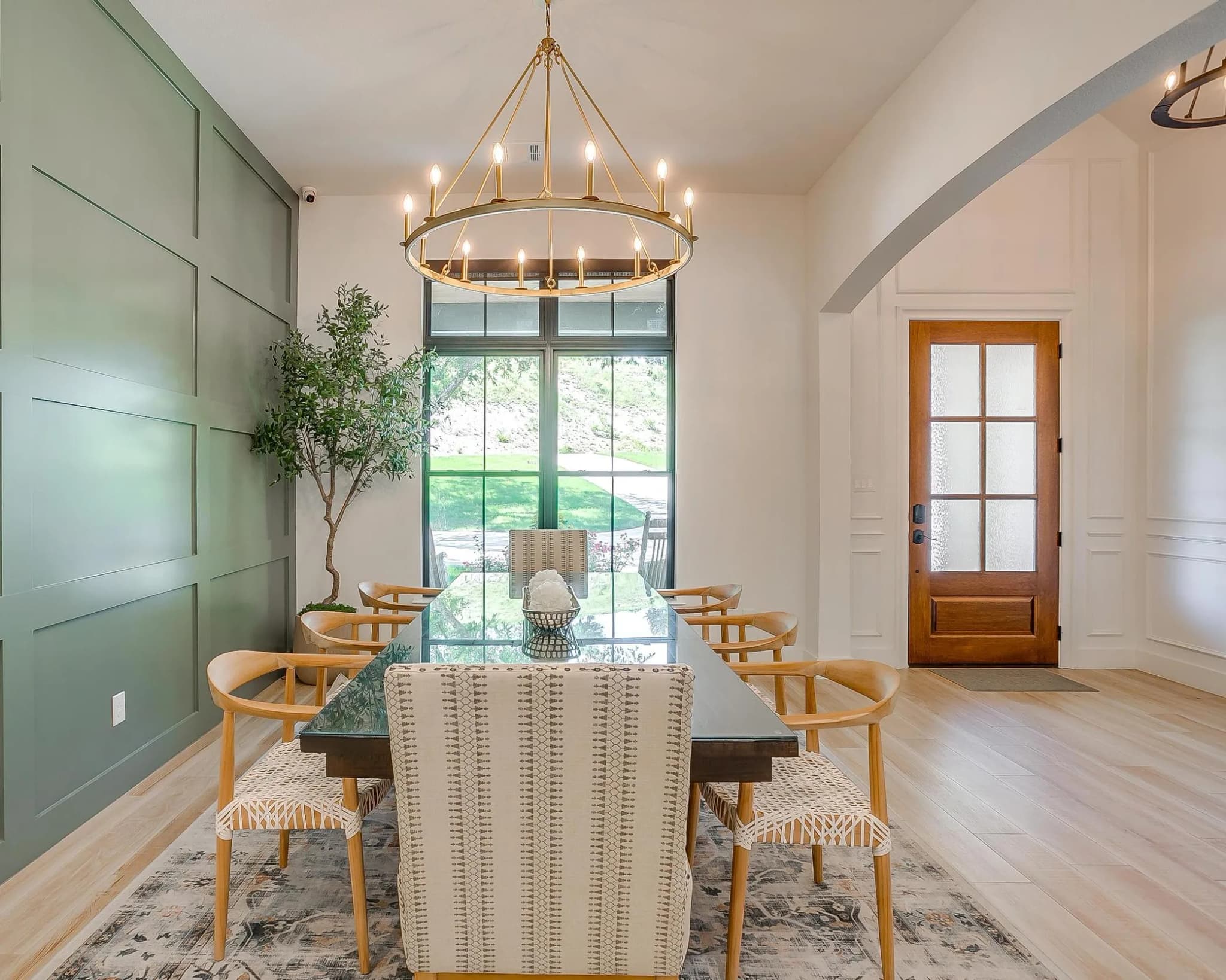 The dining room: a sage-green paneled wall behind a long table, a ring chandelier overhead, and a wood door seen through an arch.