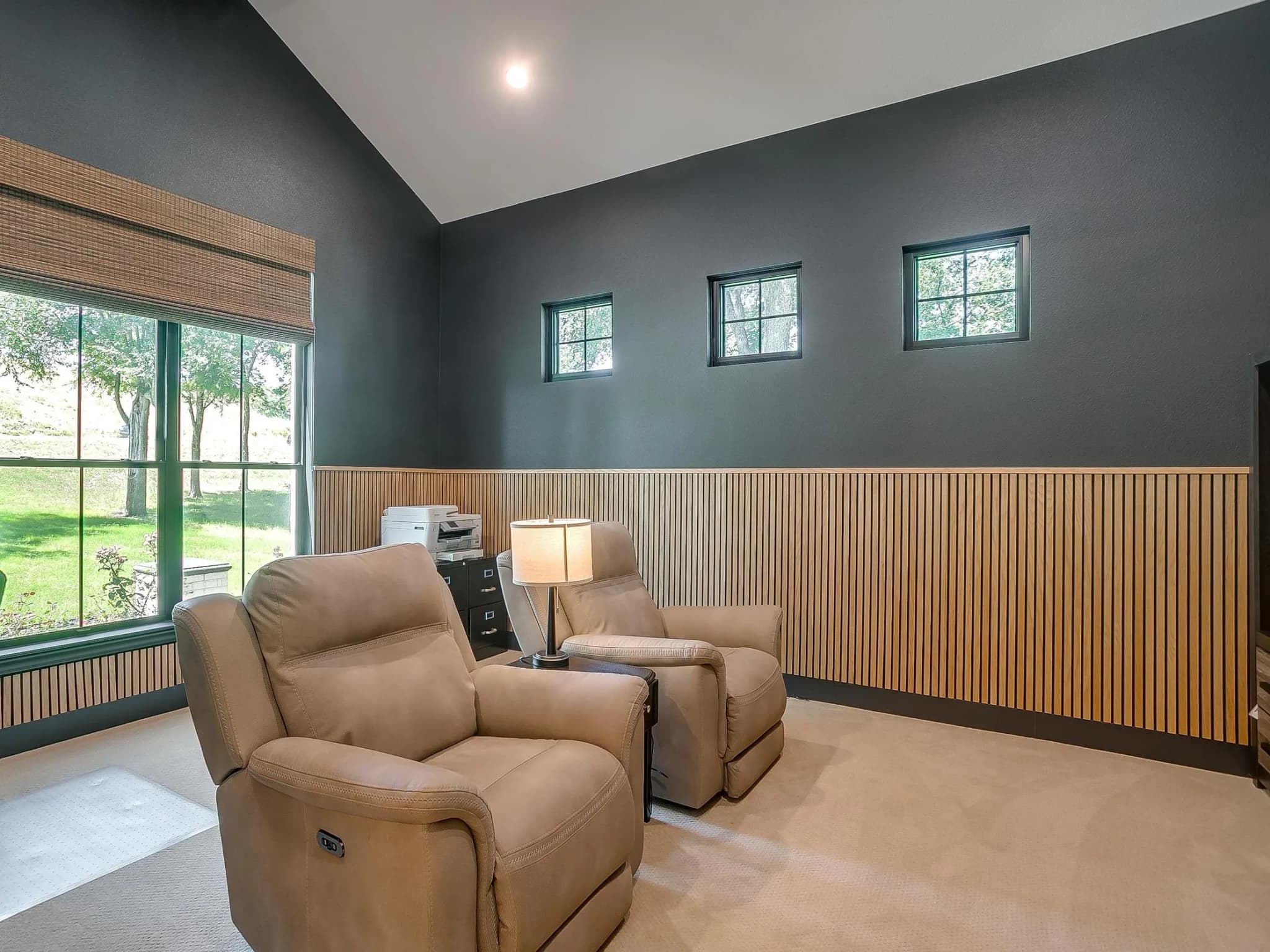 A study with dark walls, a slatted wood wainscot and two recliners facing tall windows onto the trees.