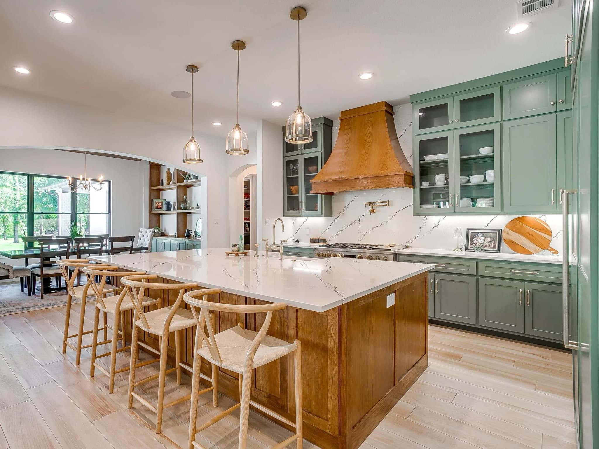 The kitchen from the breakfast room: a long wood island with counter stools, sage cabinets, glass-front uppers and a wood range hood.