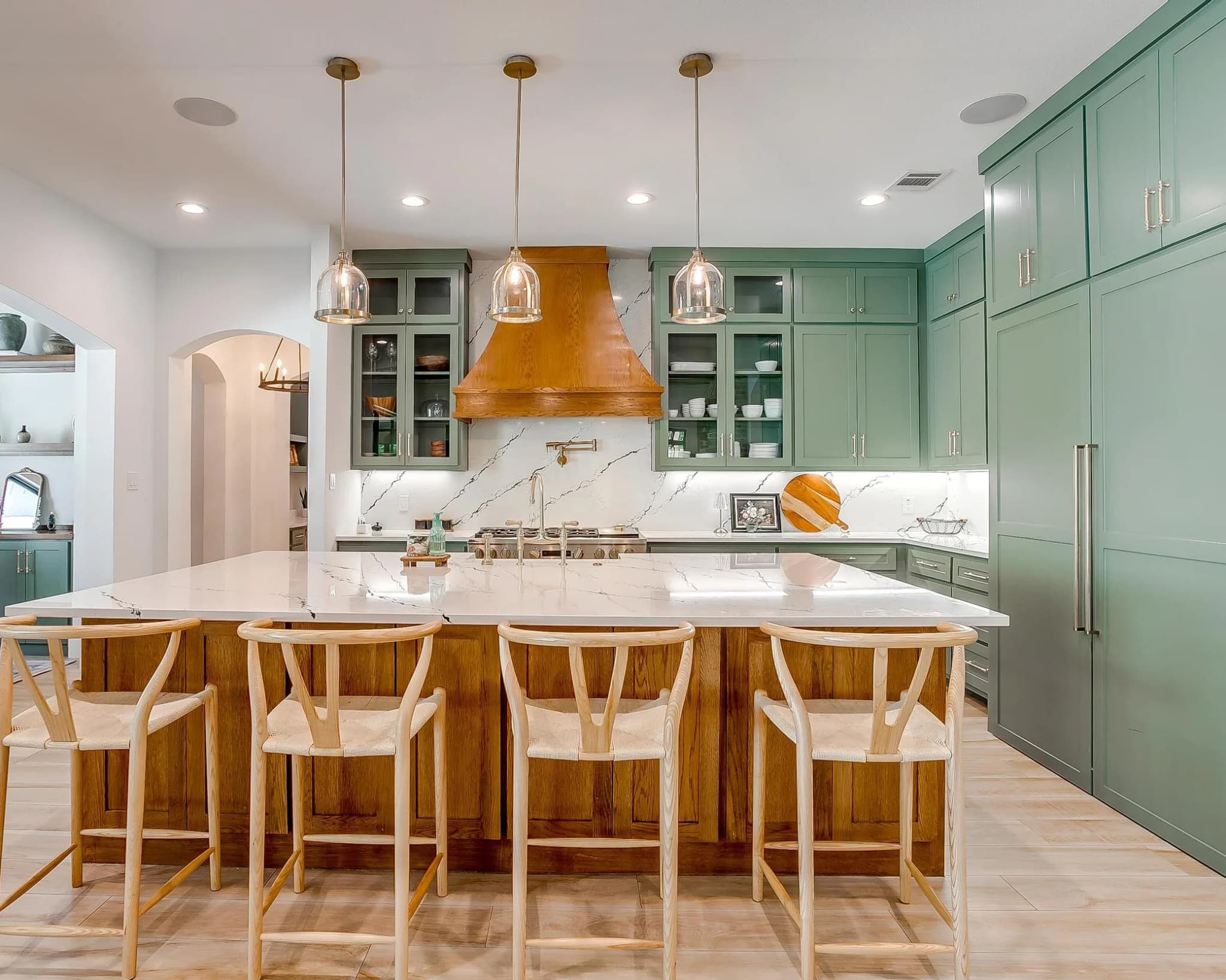 The kitchen straight on: sage cabinets to the ceiling, a wood range hood centered on a white veined backsplash, and a wood island lined with stools.