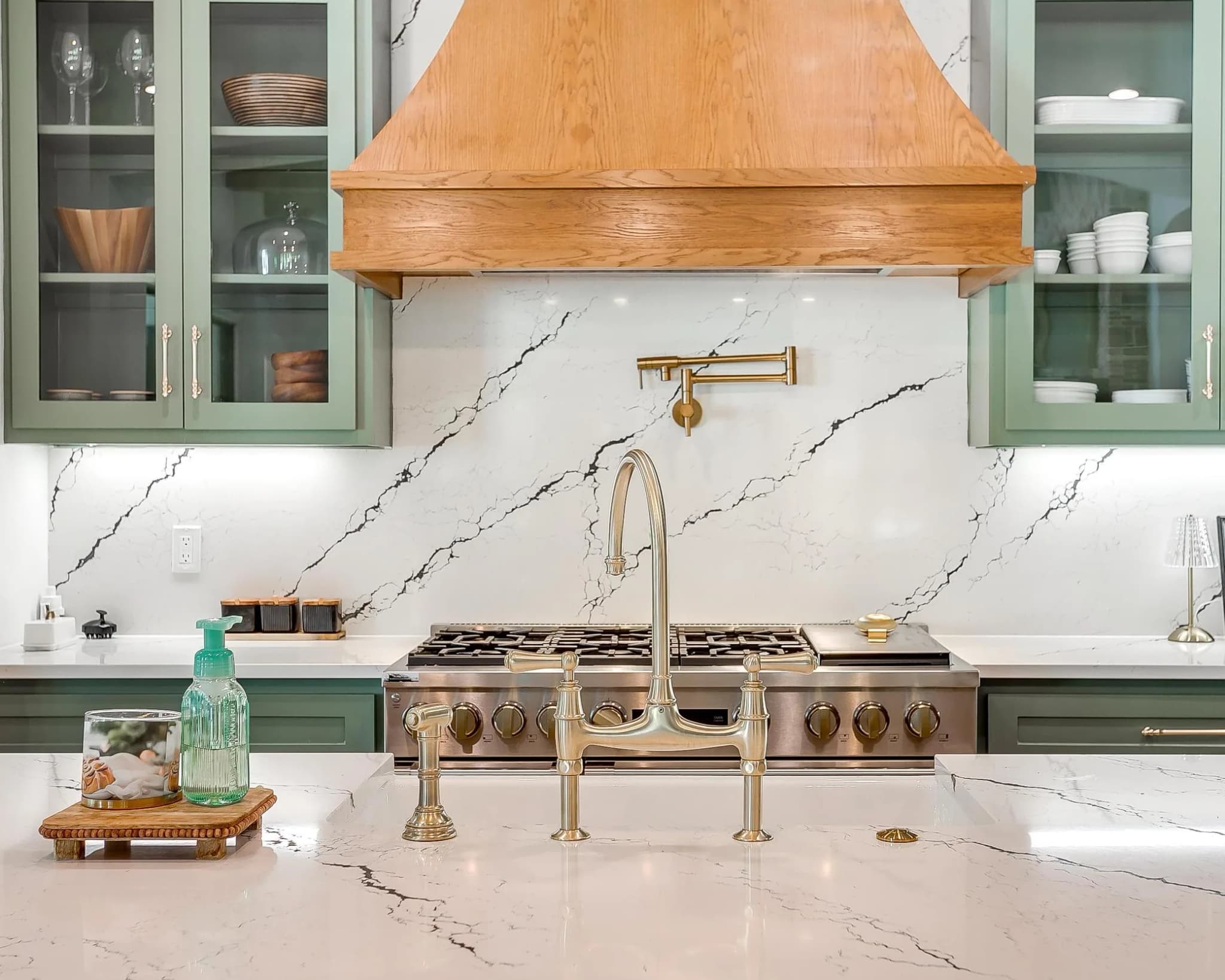 The range wall: a wood hood over a white veined slab backsplash, a brass pot filler, and glass-front cabinets to either side.