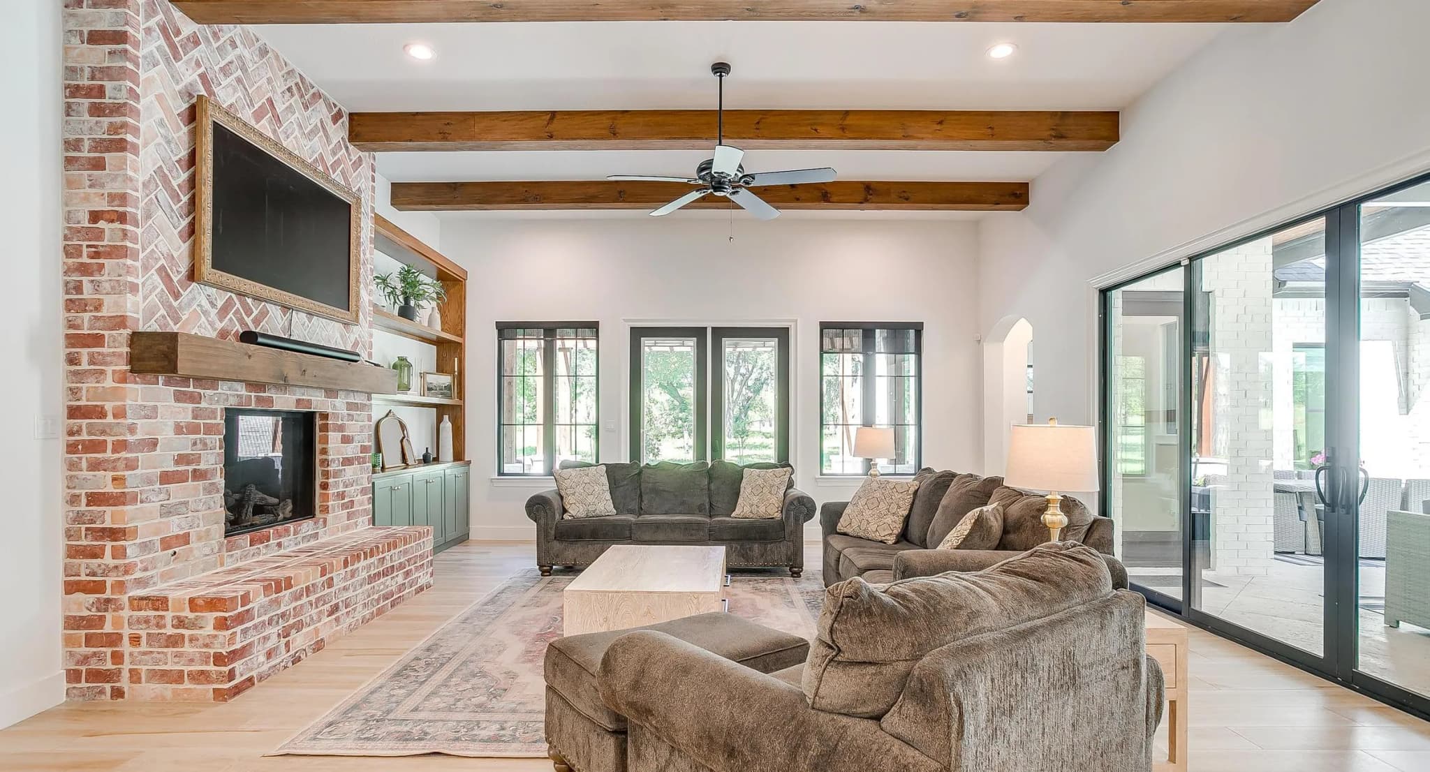 The living room: a brick fireplace with a herringbone inset and a wood mantel under exposed beams, and a wall of black-framed windows onto the patio.