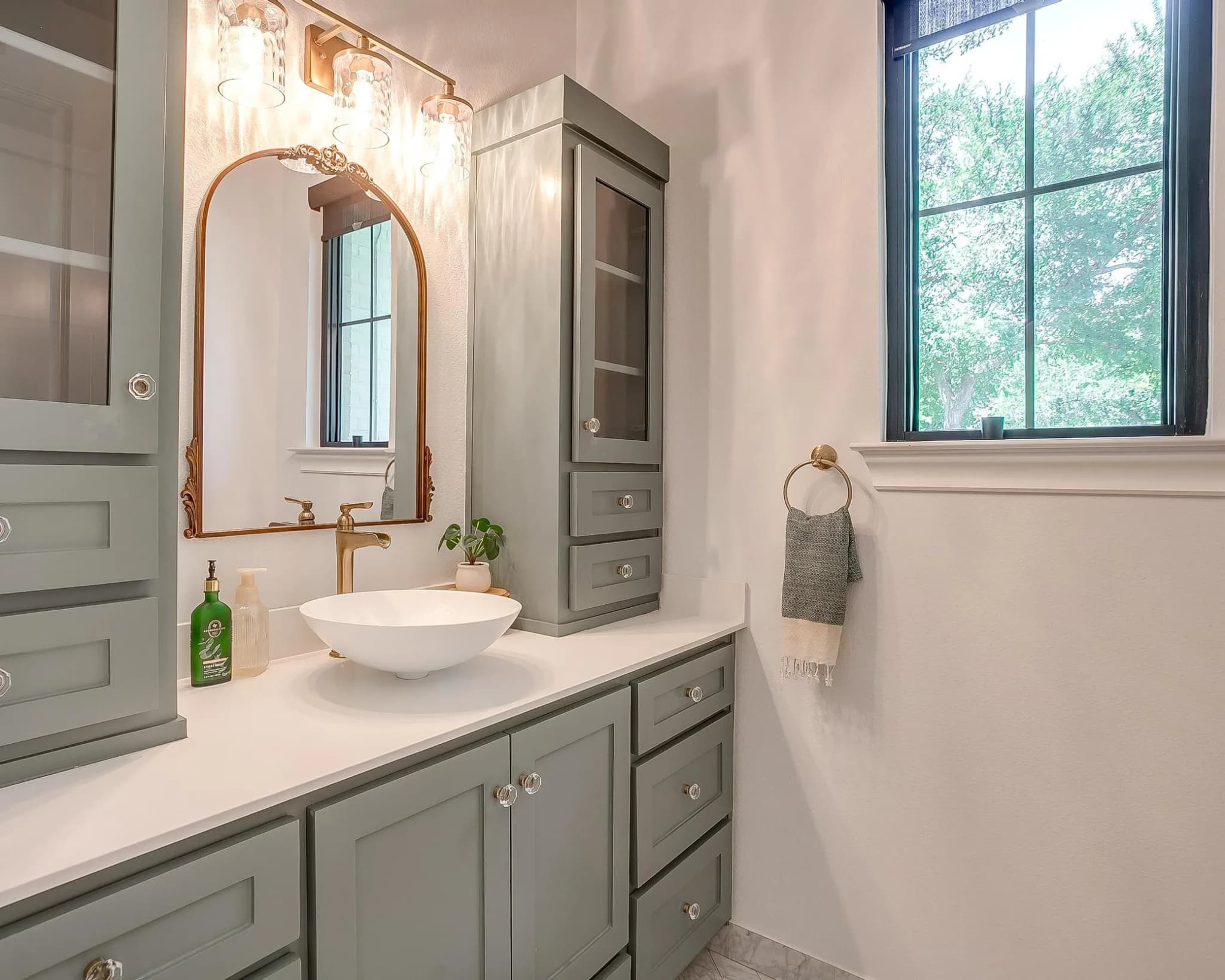 A bath vanity in sage with a vessel sink, an arched brass-framed mirror and glass-front cabinets, a black-framed window beside it.