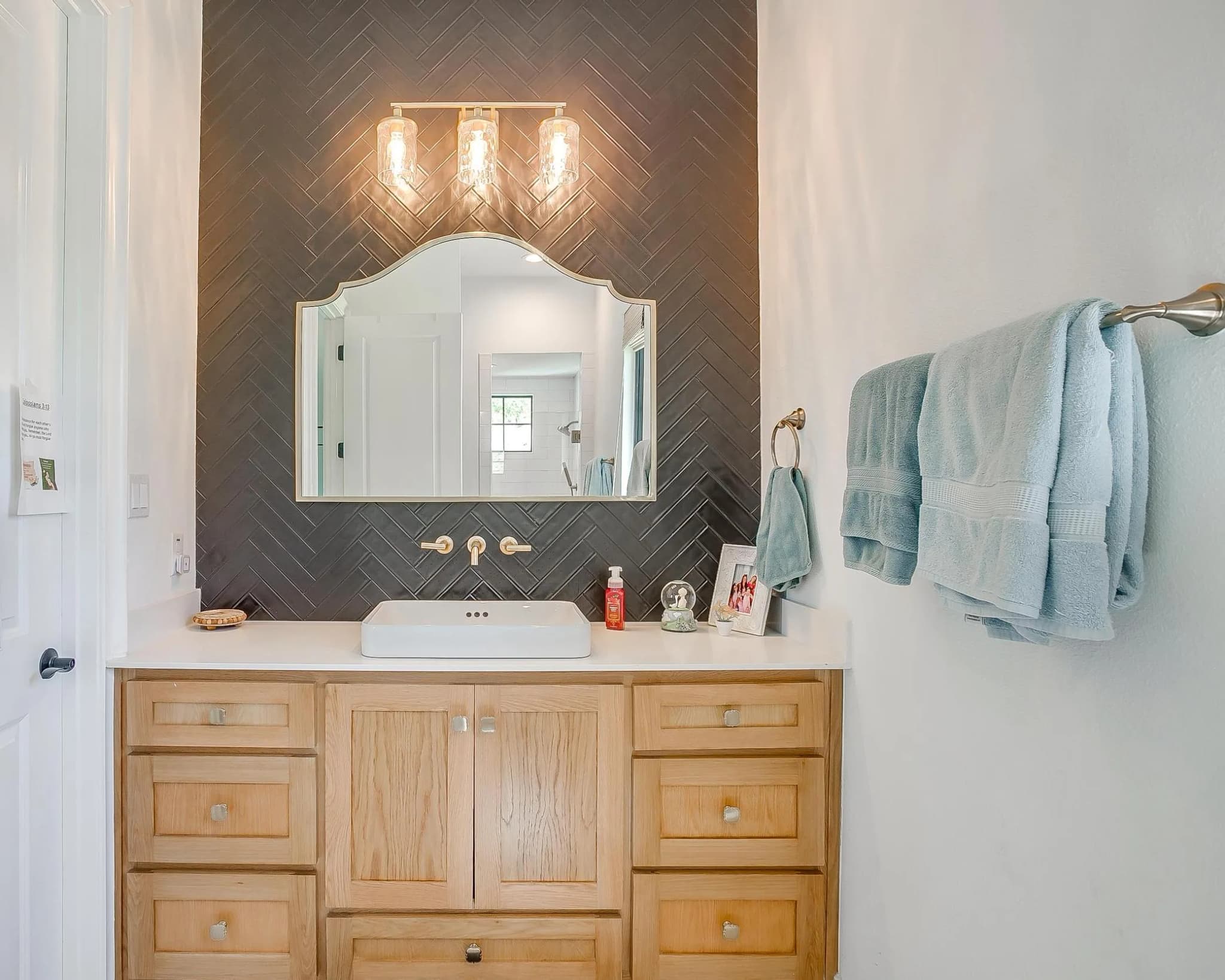 A guest bath: dark herringbone tile behind an arched mirror, a wood vanity and brass lights.