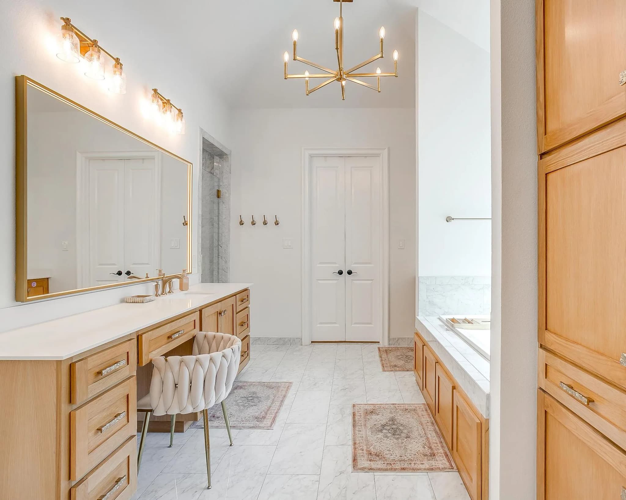 The primary bath: a long wood vanity under a brass-framed mirror, a tub at the far end, and a chandelier overhead.