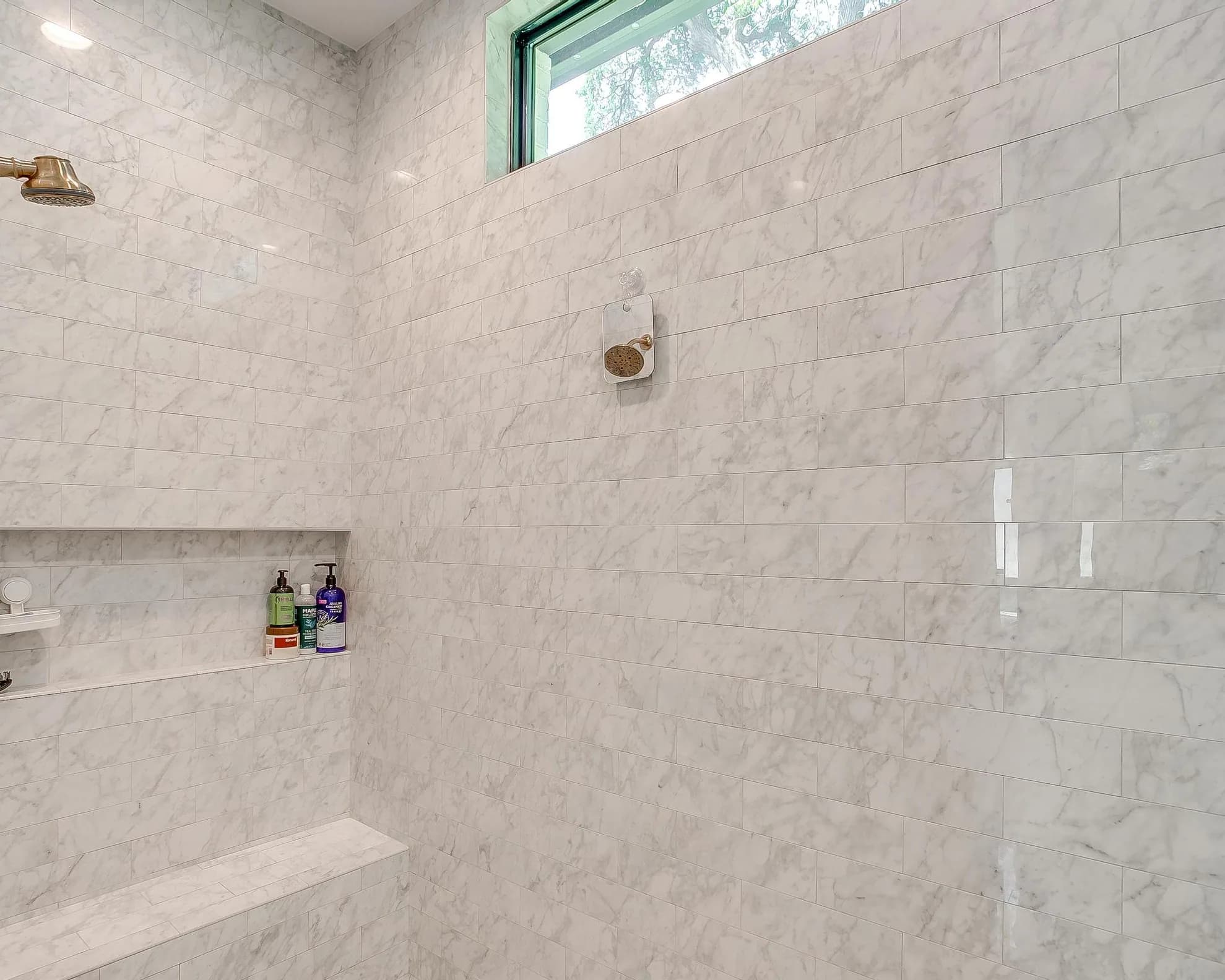 A walk-in shower in large-format white veined tile with a niche and a high window.