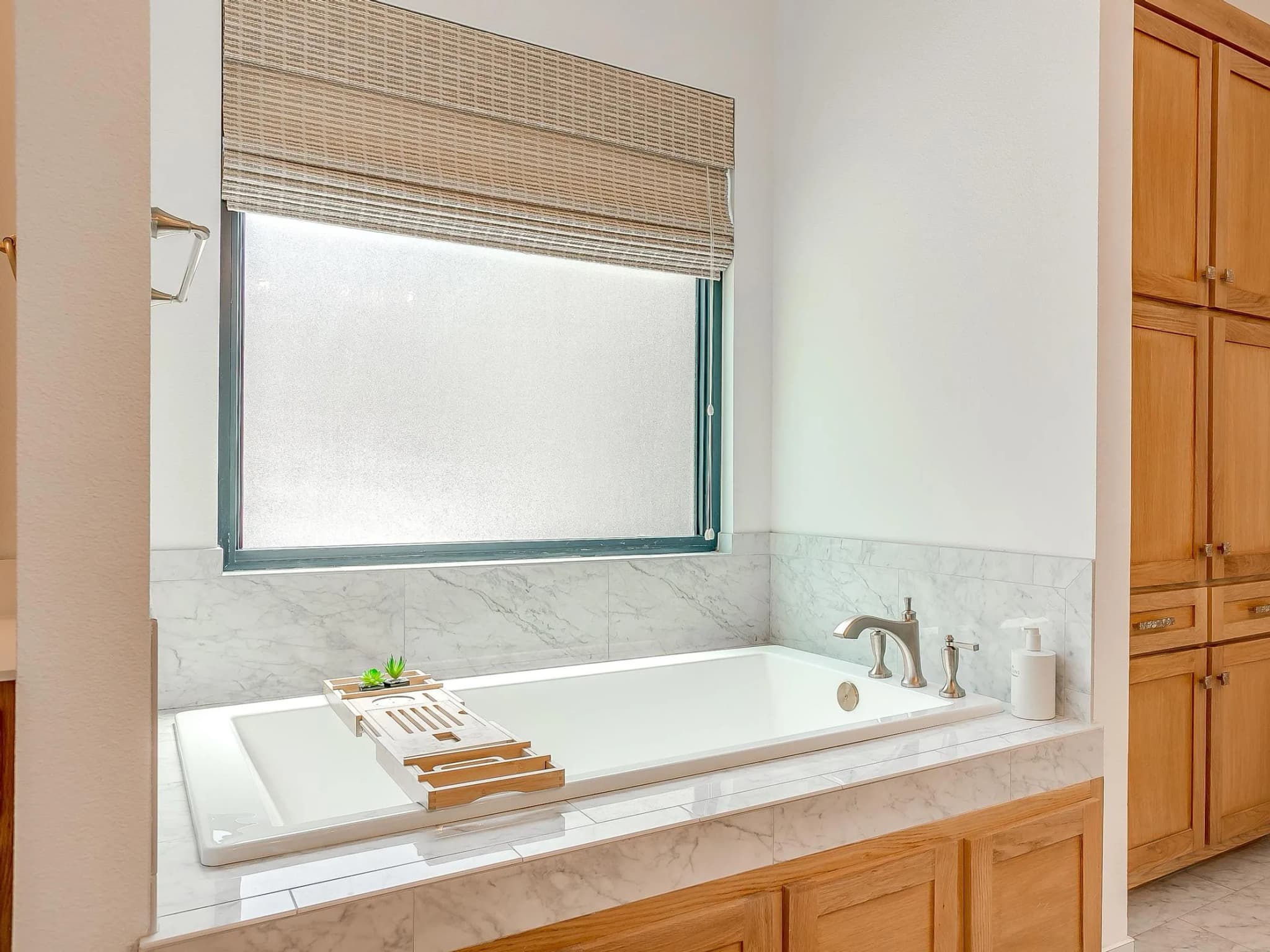 A drop-in tub under a wide window, framed in wood cabinetry and white veined tile.