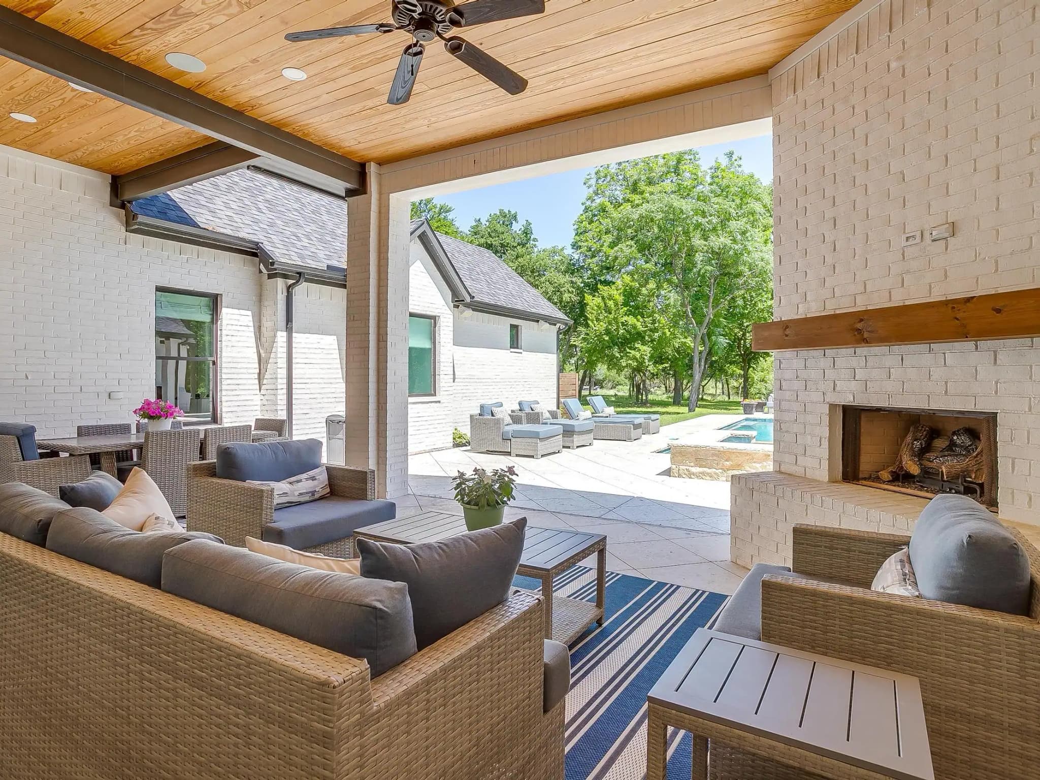Under the covered patio: a white brick fireplace with a wood mantel, a wood-planked ceiling, and seating turned toward the pool.