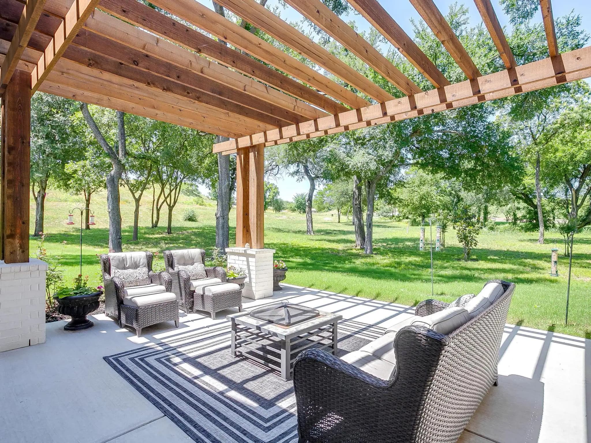 Under a wood pergola: outdoor seating on a striped rug, looking out to the lawn and trees.