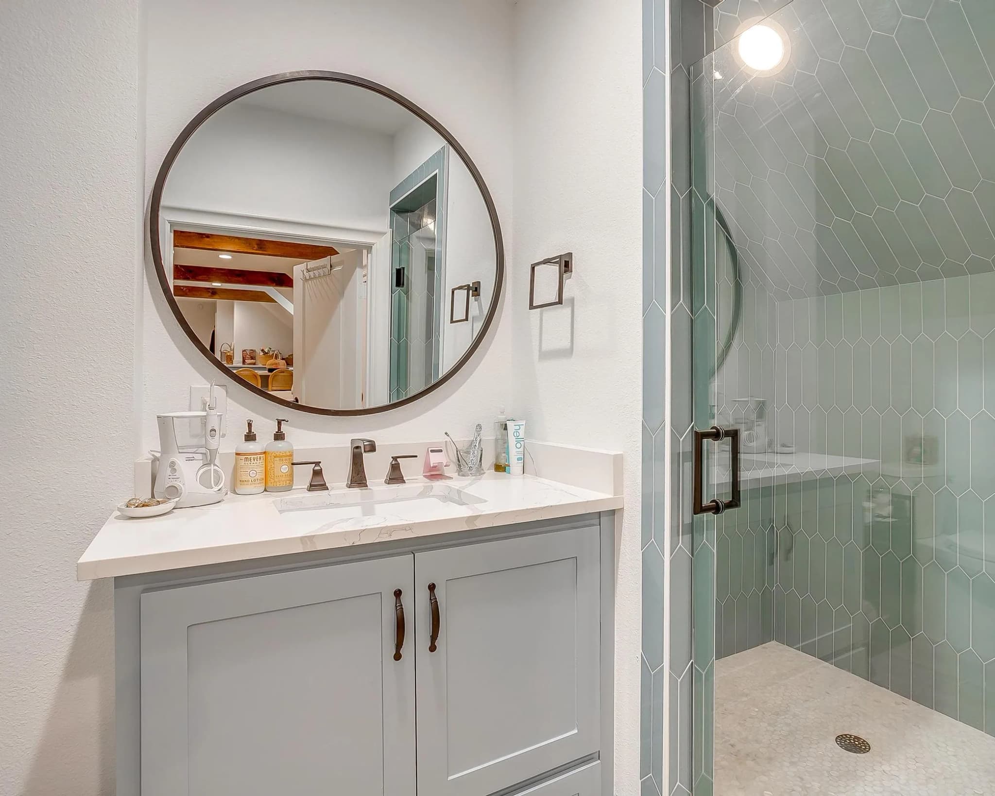 The garage-room bath: a round mirror over a gray vanity and a shower in pale green elongated tile.