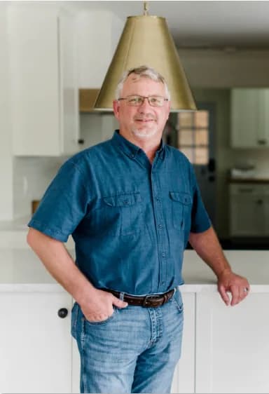 A man with gray hair and glasses in a blue short-sleeved denim shirt, one hand in his pocket, leaning against a white kitchen island under a brass pendant.