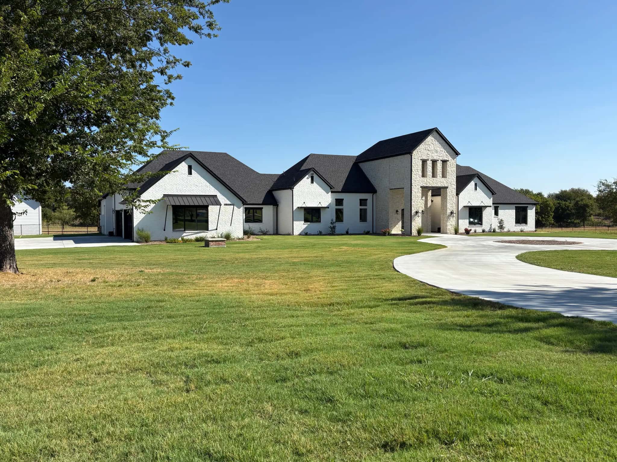 A single-story white brick house with black roofs, a stone entry tower with three slot windows, and a circular drive across a new lawn under a big sky.