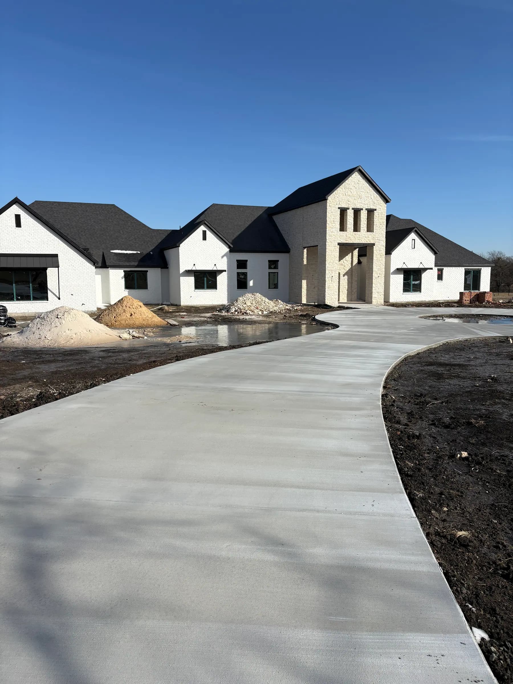 The same house before landscaping: a fresh concrete drive curving to the stone entry, piles of sand and rock, bare ground under a pale sky.