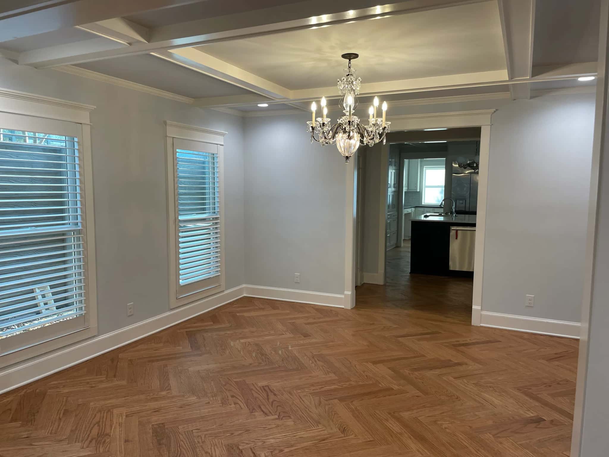 A dining room with a coffered ceiling and a glass-drop chandelier, tall cased windows with wood blinds, opening to a kitchen with dark cabinets.