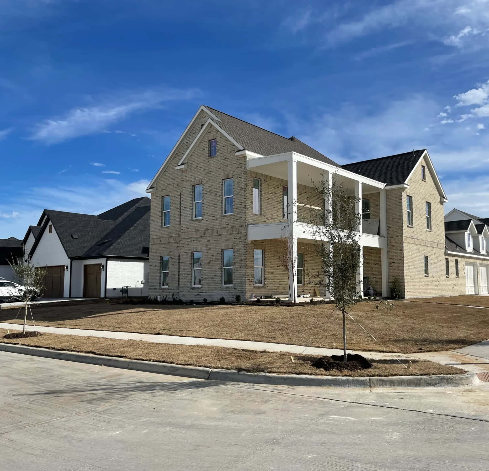 A corner-lot two-story house in pale blond brick with a double gallery porch on square white columns, newly sodded and planted with staked trees.