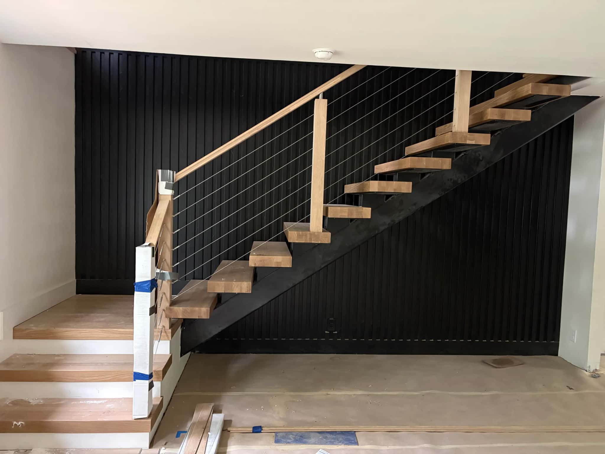 A floating pale wood stair on a black steel stringer with cable rails, against a wall of black vertical slats, the treads still taped for protection.