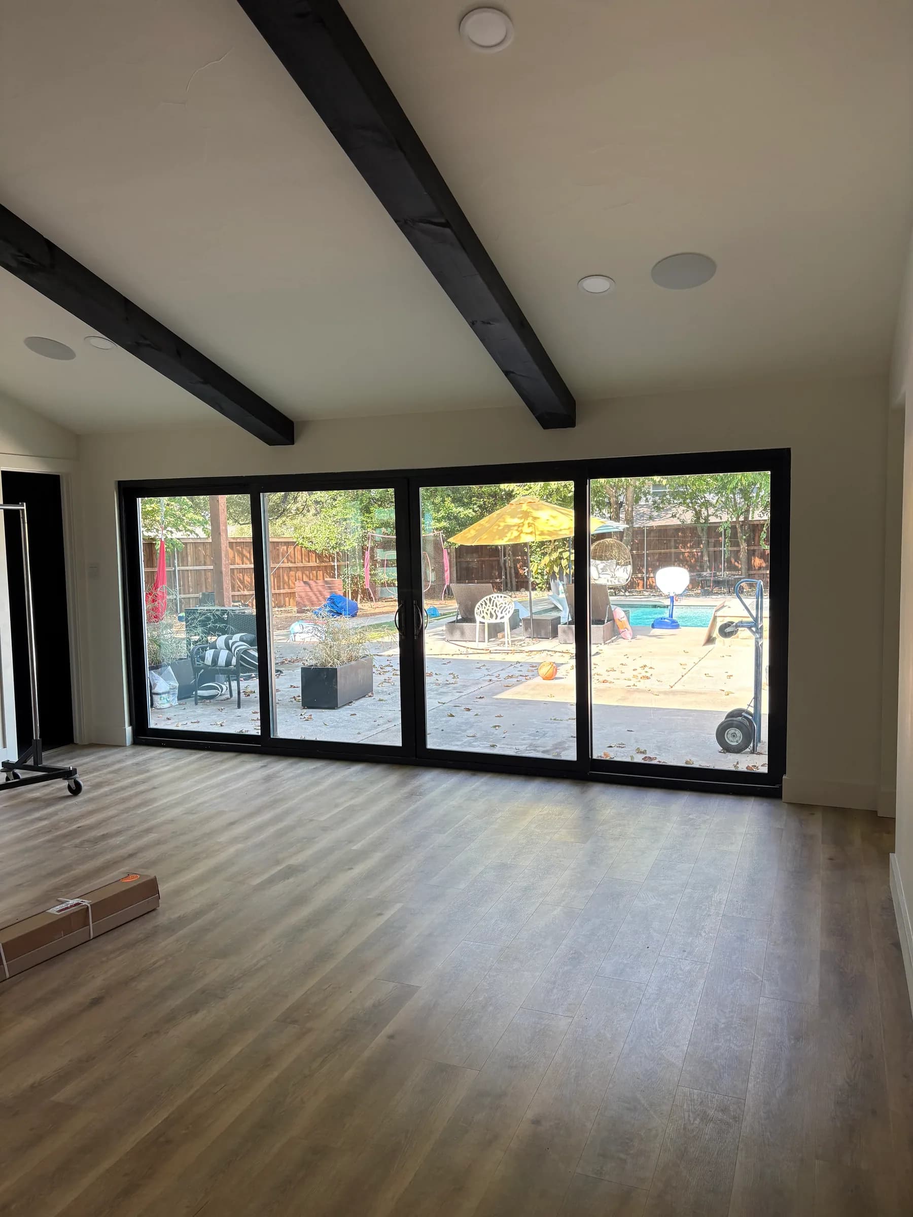 A vaulted room with dark beams and a wall of black-framed sliding doors onto the pool terrace, wood floors just laid.