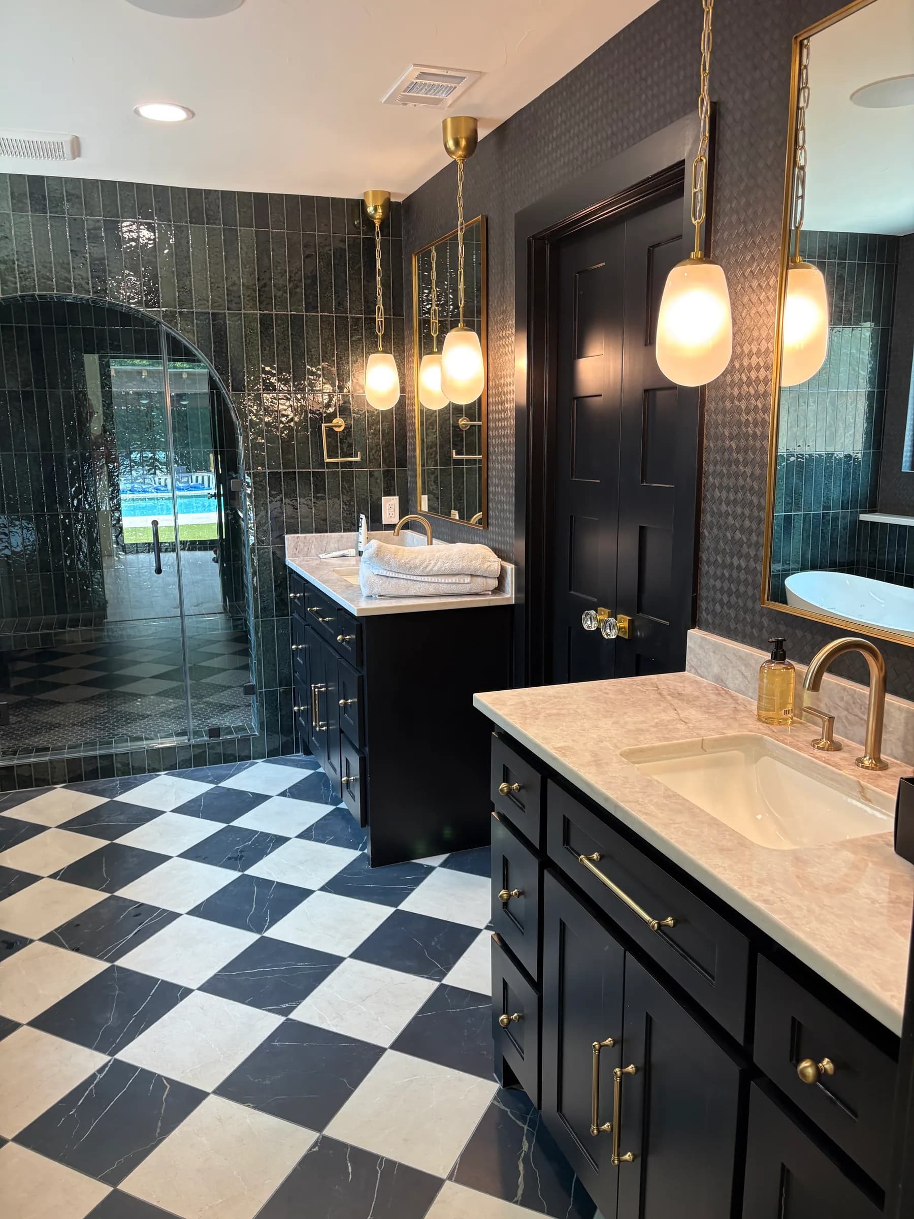 A bathroom in glossy black tile with a checkerboard stone floor, an arched glass shower door, a black vanity with brass pulls and globe pendants.