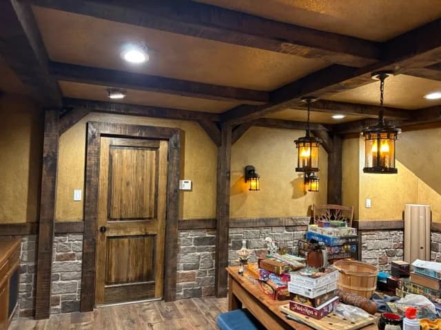 A game room finished like a tavern: heavy timber beams and posts, textured ochre walls over a stone wainscot, lantern sconces and a plank door.