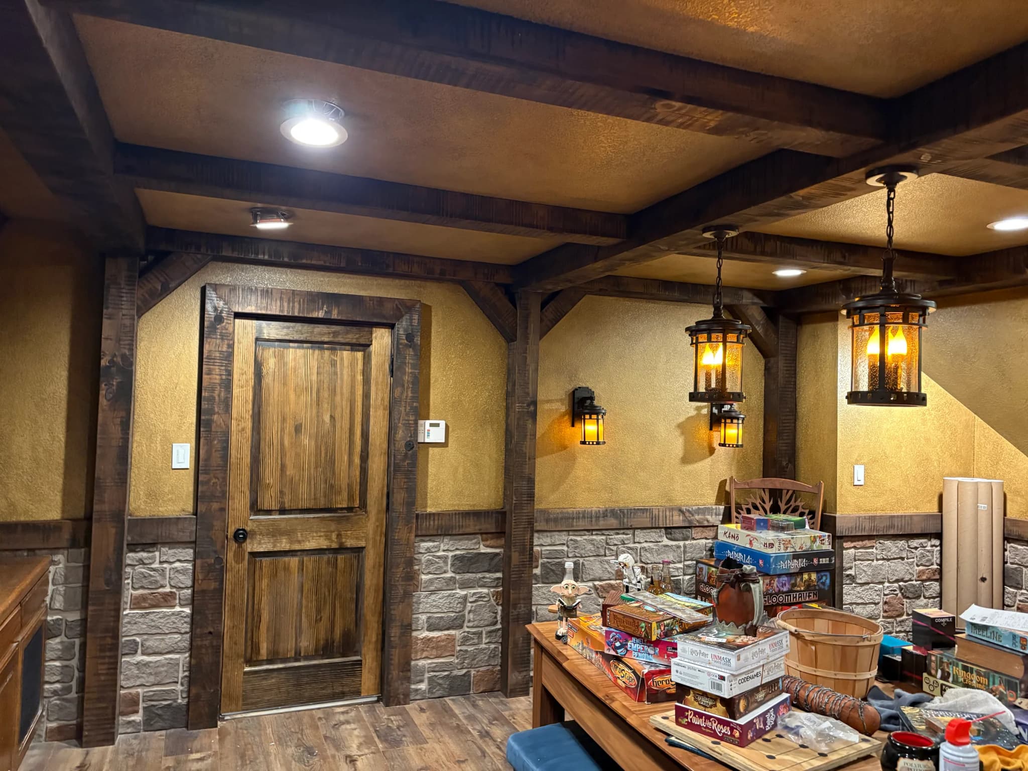 The game room's beamed ceiling and lantern pendants over a heavy timber table, with the stone wainscot along the stair wall.