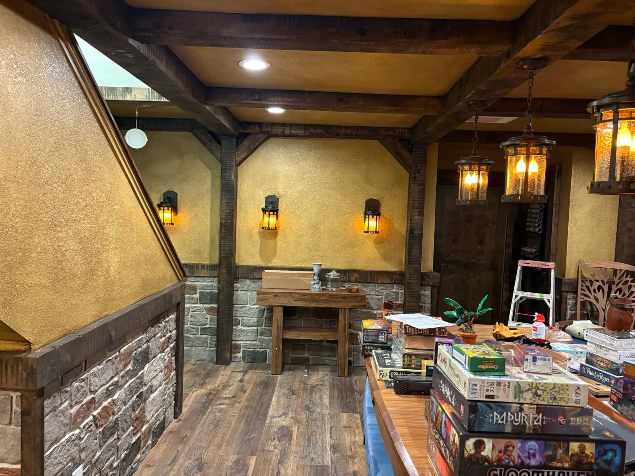 A game room finished like a tavern: heavy timber beams and posts, textured ochre walls over a stone wainscot, lantern pendants and a plank floor.