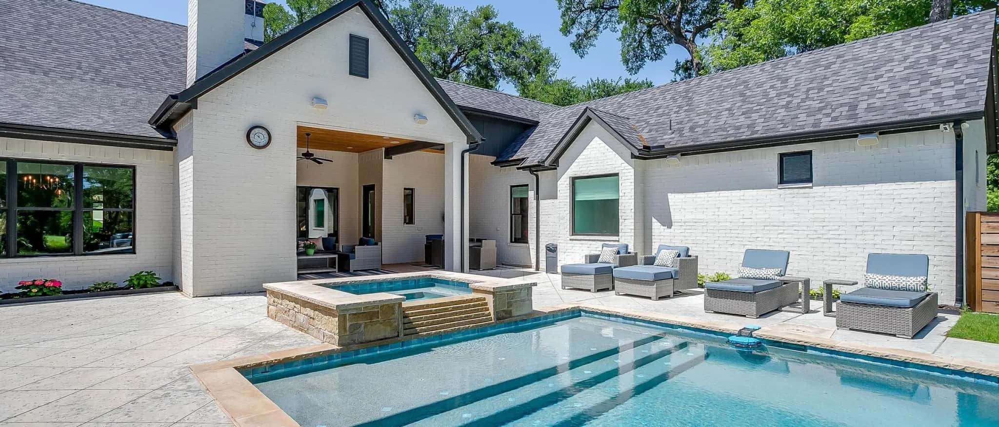 The back of a single-story white brick house with a tall chimney and steep gables, its covered patio open to a pool with a raised spa and wide steps into the water.