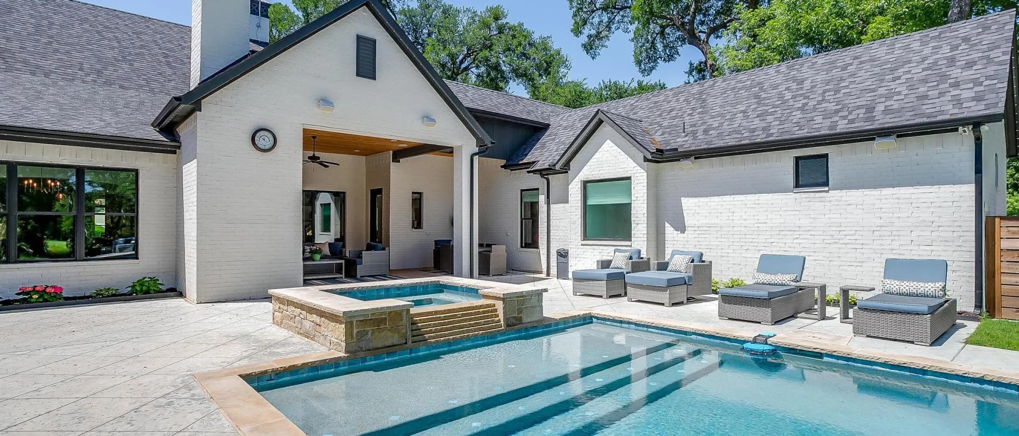 The back of a single-story white brick house with a tall chimney and steep gables, its covered patio open to a pool with a raised spa and wide steps into the water.