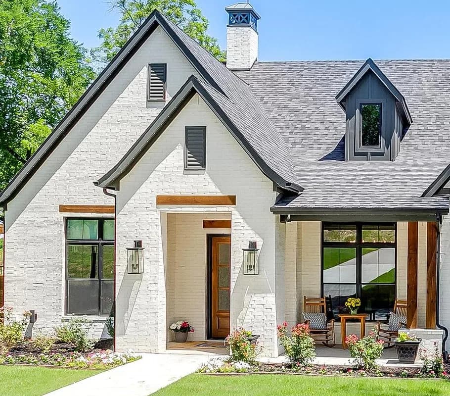 The entry gables in white brick, a wood door under a timber lintel between two lanterns, and a covered porch with rocking chairs beside it.